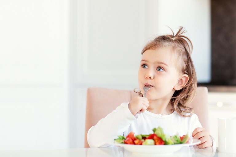 Child girl eating fresh raw vegetables vitamin salad in white kitchen background and thoughtfully looks at the empty copy space.Healthy nutrition food for children. Place for text for advertising.